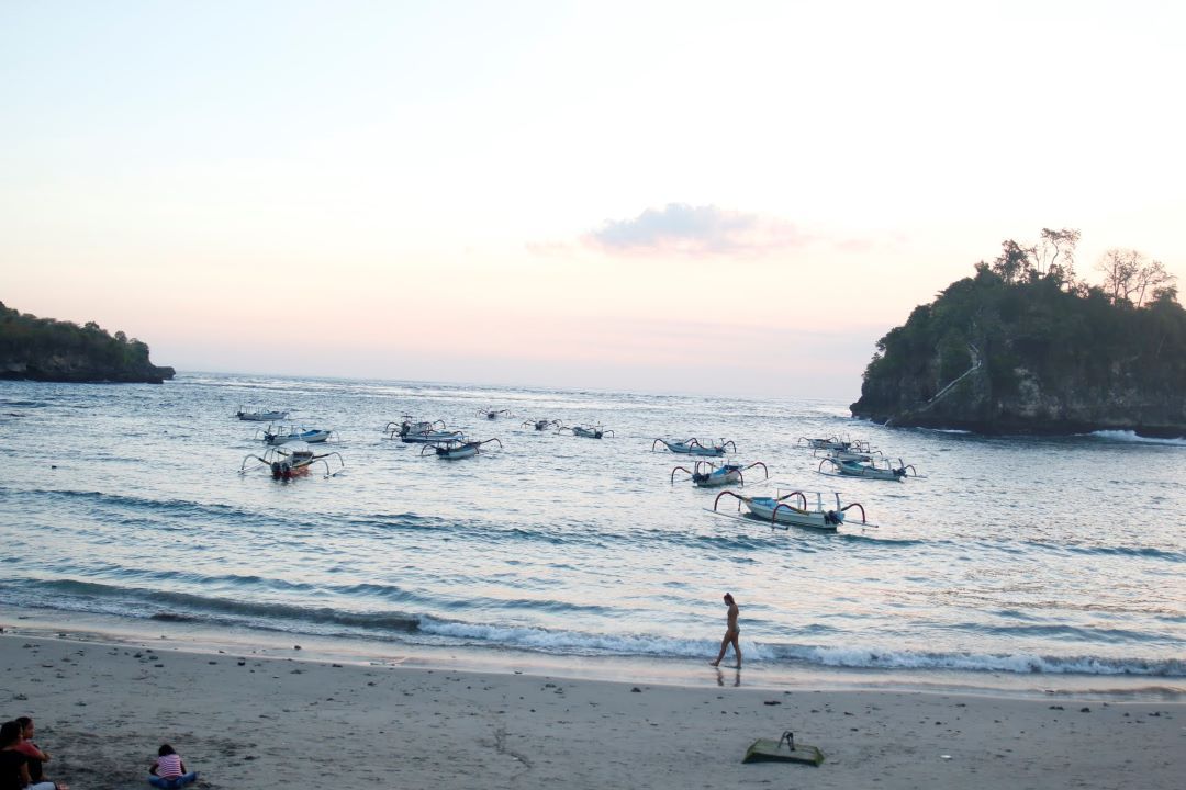 ruta por indonesia vistas de playa al atardecer con pequeñas barcas en el agua y chica caminando por la orilla